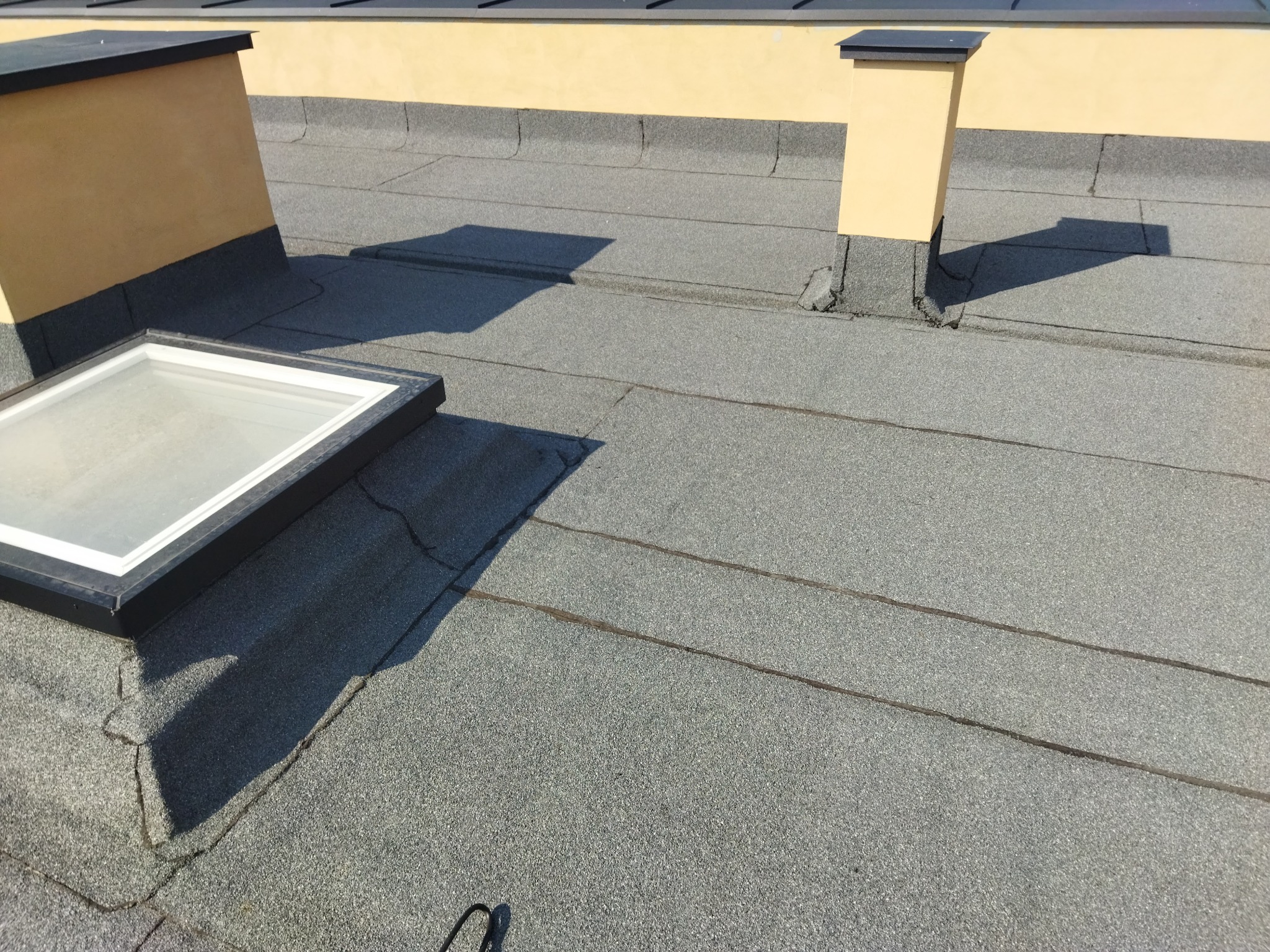 Płaski dach pokryty papą z widocznym świetlikiem dachowym i kominem wentylacyjnym, na tle żółtej ściany budynku.