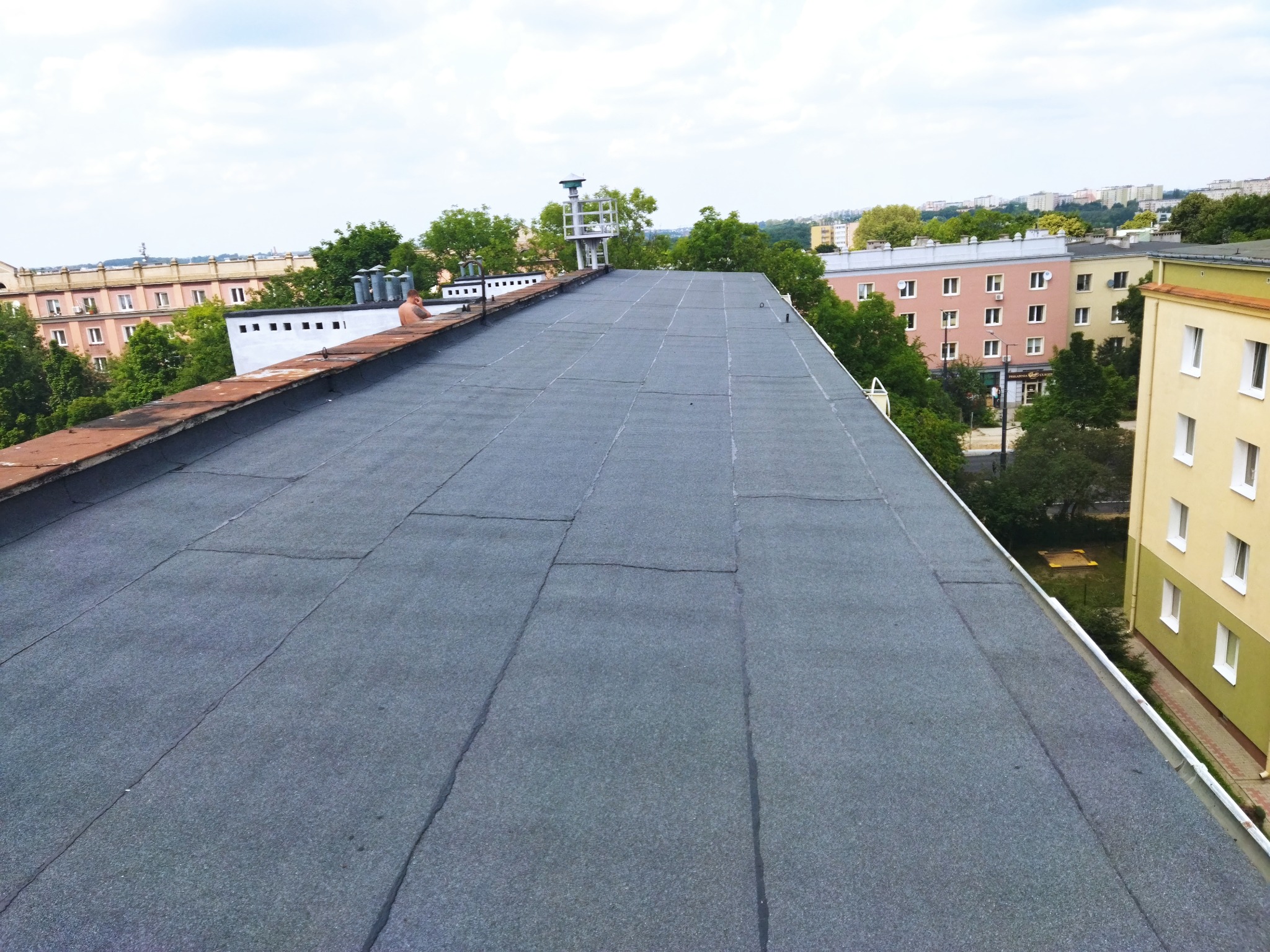 Płaski dach pokryty szarą papą termozgrzewalną na budynku wielorodzinnym, widok z góry na miasto w tle, widoczny fragment instalacji odgromowej.