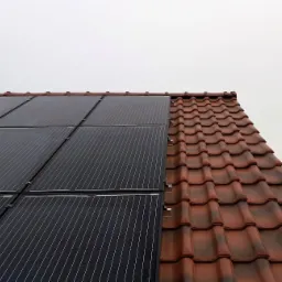 Częściowy widok instalacji paneli słonecznych na dachu z czerwoną dachówką, z anteną w tle i pochmurnym niebem.