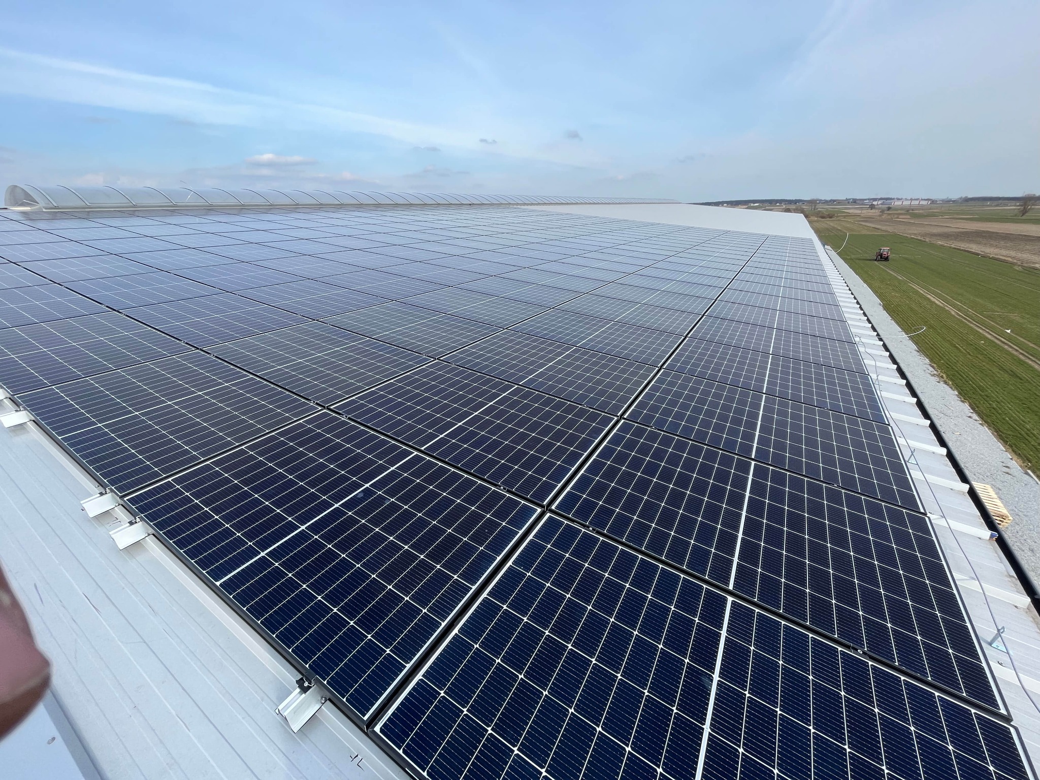 Rozległa instalacja paneli słonecznych na dachu hali przemysłowej, widok z góry, w tle pola uprawne i niebo z lekkimi chmurami.