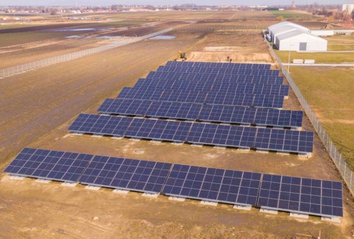 Rozległa instalacja paneli słonecznych na betonowych podstawach, widok z góry na brązowe pole z budynkiem magazynowym w tle.