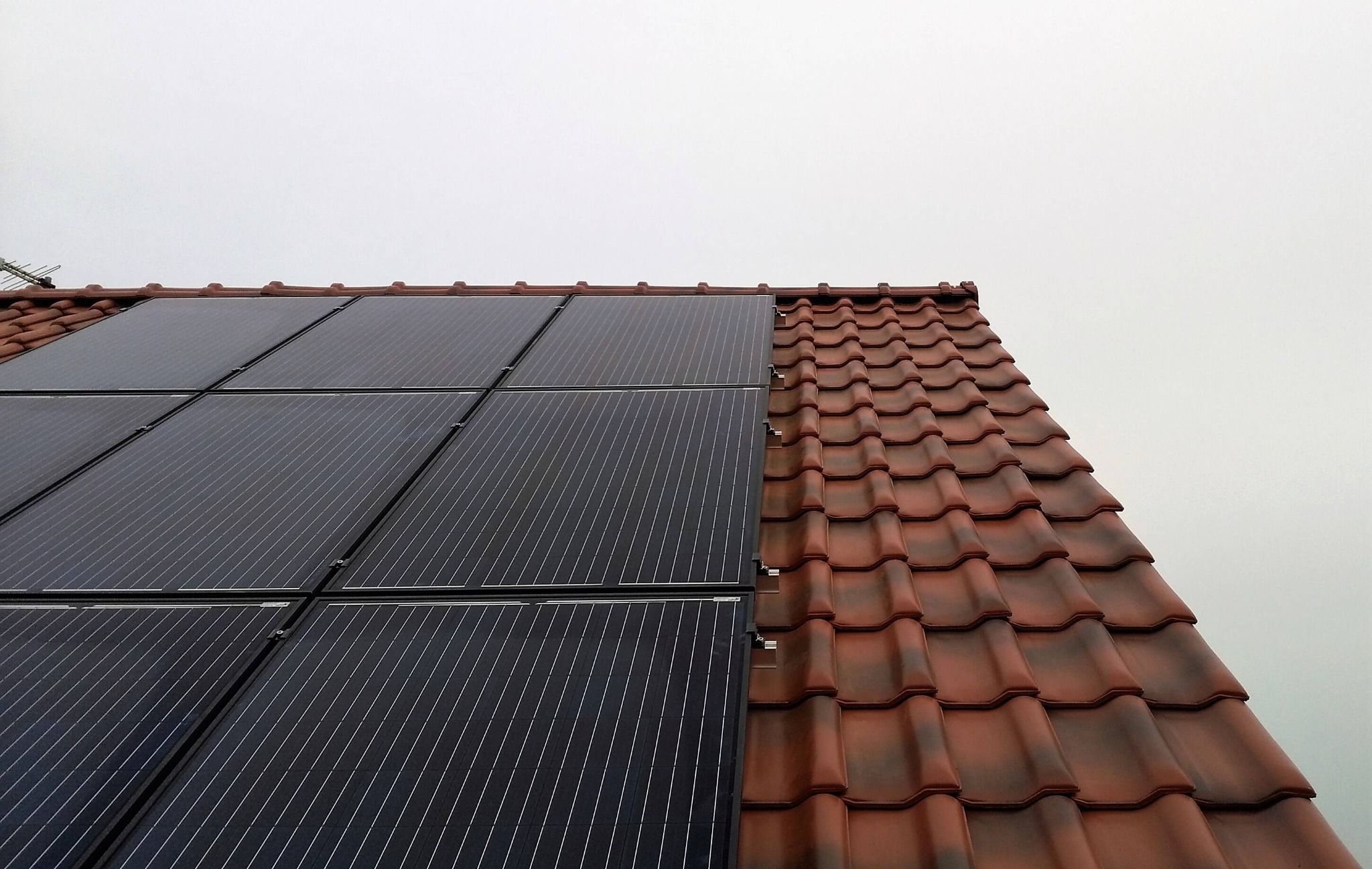 Częściowy widok instalacji paneli słonecznych na dachu z czerwoną dachówką, z anteną w tle i pochmurnym niebem.