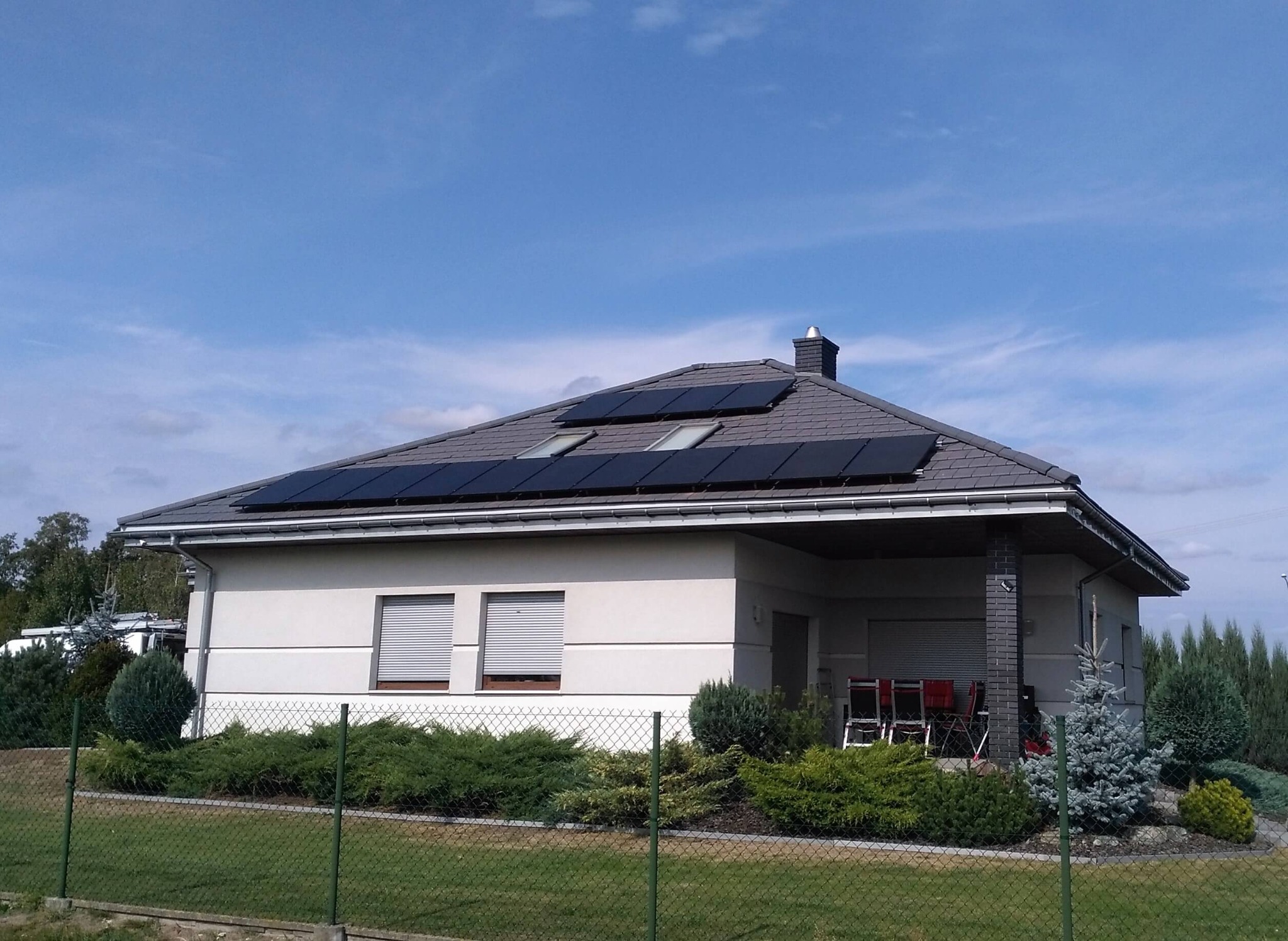 Jednorodzinny dom z szarą dachówką, na której zamontowano czarne panele słoneczne. Widoczne okna dachowe i starannie wypielęgnowany ogród.