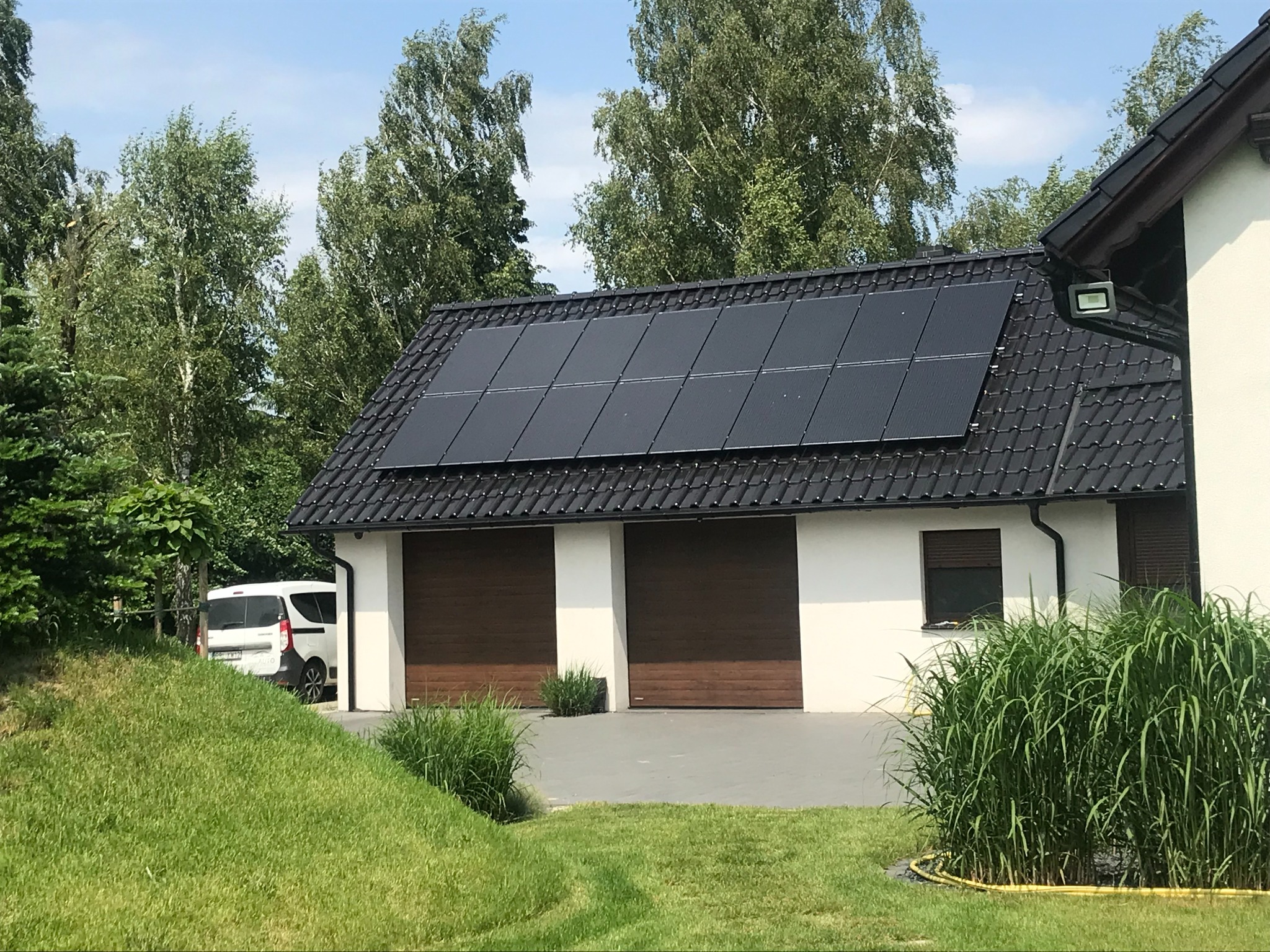 Czarny dach dwuspadowy z panelami słonecznymi nad garażem z dwoma brązowymi bramami, na podjeździe zaparkowany biały samochód dostawczy.