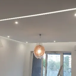 Nowoczesne oświetlenie sufitowe z ledowym paskiem i trzema drewnianymi lampami kulistymi w folii ochronnej, klimatyzator na ścianie, widok na zewnątrz przez przeszklone drzwi balkonowe.