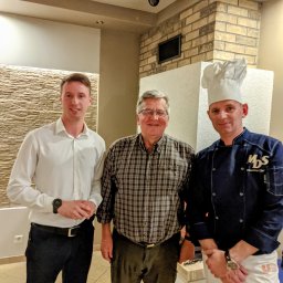 Restauracja MDS - Trzech mężczyzn pozuje do zdjęcia w restauracji, kucharz w uniformie i wysokiej czapce stoi obok klienta, w tle widoczne kamery monitoringu.