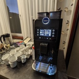 Restauracja MDS - Profesjonalny, czarny ekspres do kawy Carimali z podświetlanym panelem dotykowym, otoczony stosem białych filiżanek i szklanek na ciemnym blacie, widoczny czajnik elektryczny i puszka z herbatami...