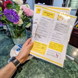 Restauracja MDS - Menu restauracji 'Restauracja MDS' w przezroczystej okładce, trzymane w dłoni, z bukietem kolorowych astra w szklanym wazonie w tle, leżące na zielonym marmurowym blacie.