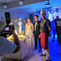 Restauracja MDS - Uroczyste przyjęcie weselne: para młoda pozuje obok szefa kuchni w czapce i czerwonym fartuchu, w tle goście, zespół muzyczny na scenie i stół z dekoracjami i fontannami iskier, filmowane przez...