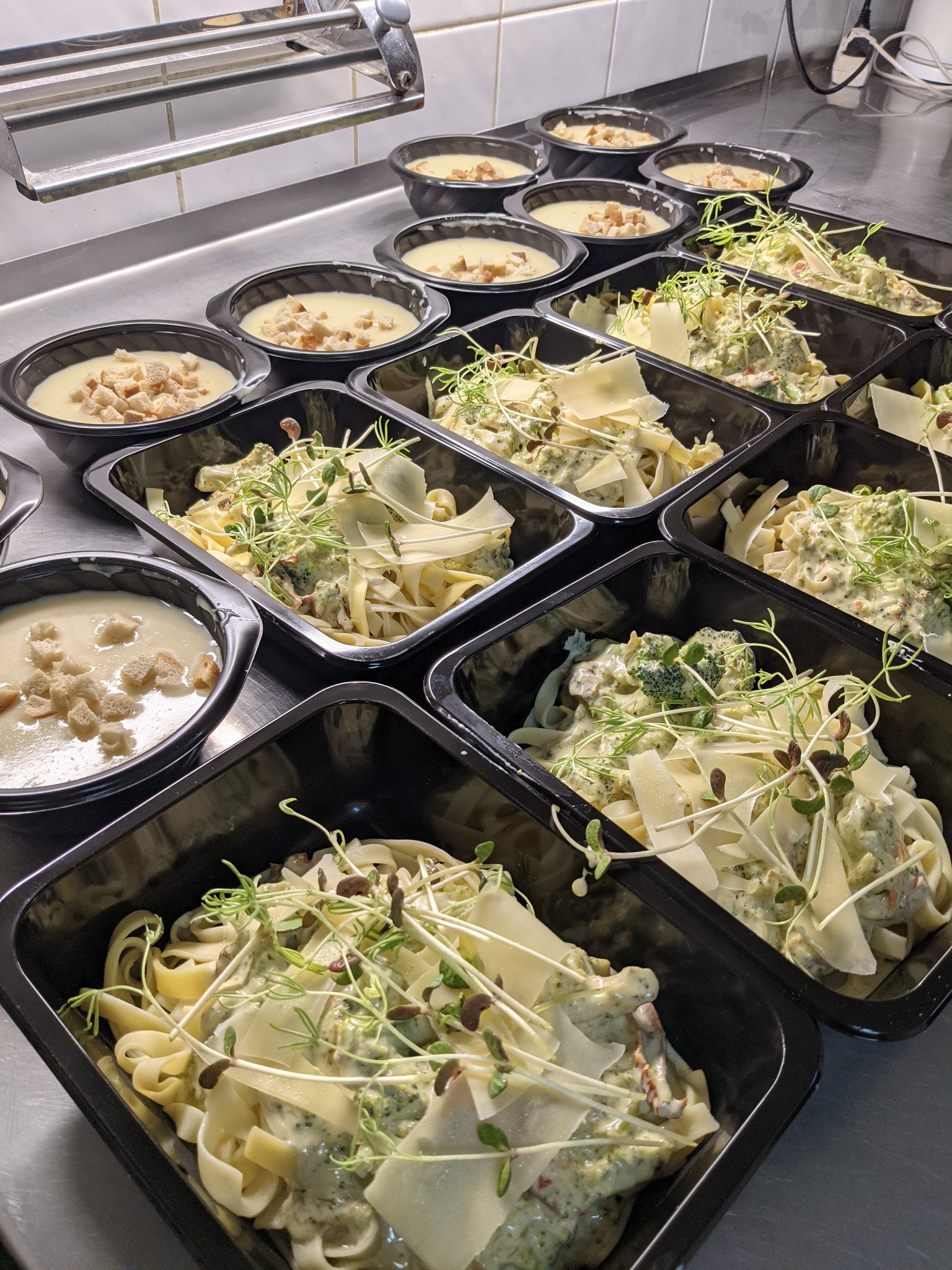Gotowe dania w czarnych pojemnikach: krem z grzankami i makaron tagliatelle z sosem pesto, brokułami, parmezanem i kiełkami, ułożone rzędami na metalowym stole.