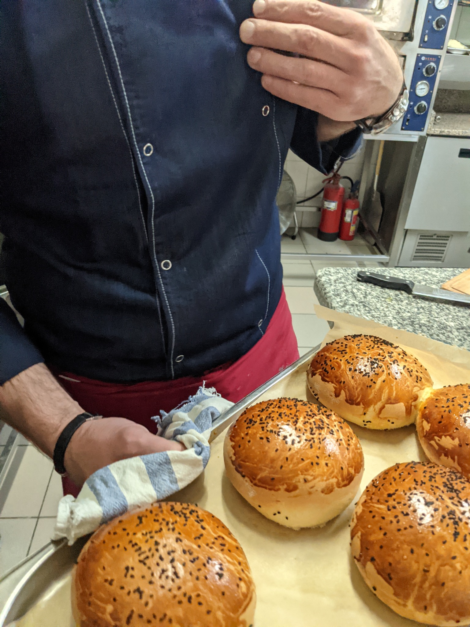 Kucharz w niebieskiej bluzie i czerwonym fartuchu, trzymający blachę z sześcioma świeżo upieczonymi, okrągłymi bułkami posypanymi czarnym sezamem, na tle profesjonalnej kuchni z piecem i czerwonymi...