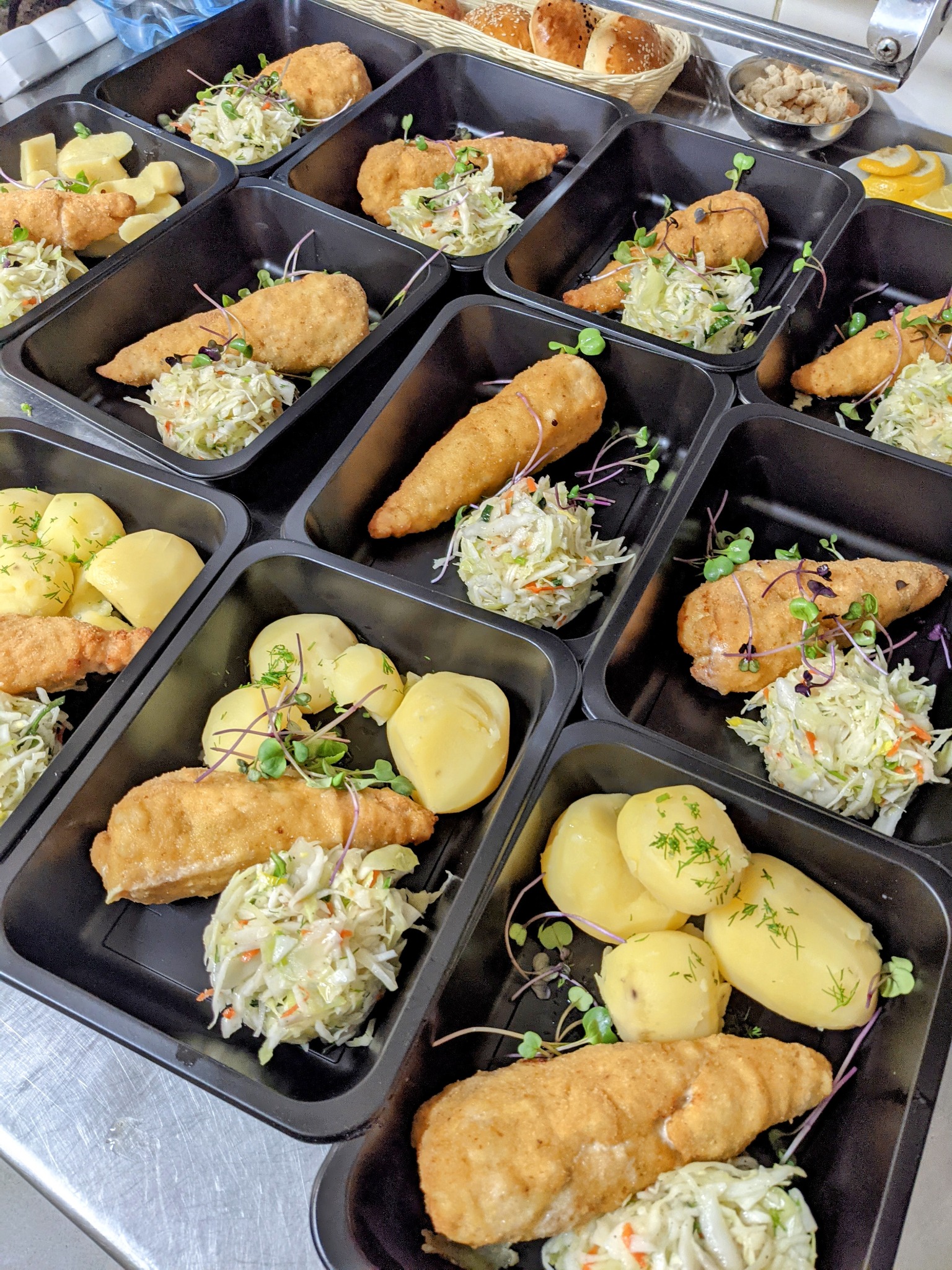 Gotowe porcje obiadowe w czarnych pojemnikach: panierowana ryba, gotowane ziemniaki posypane koperkiem oraz surówka z białej kapusty i marchewki, udekorowane microgreens.
