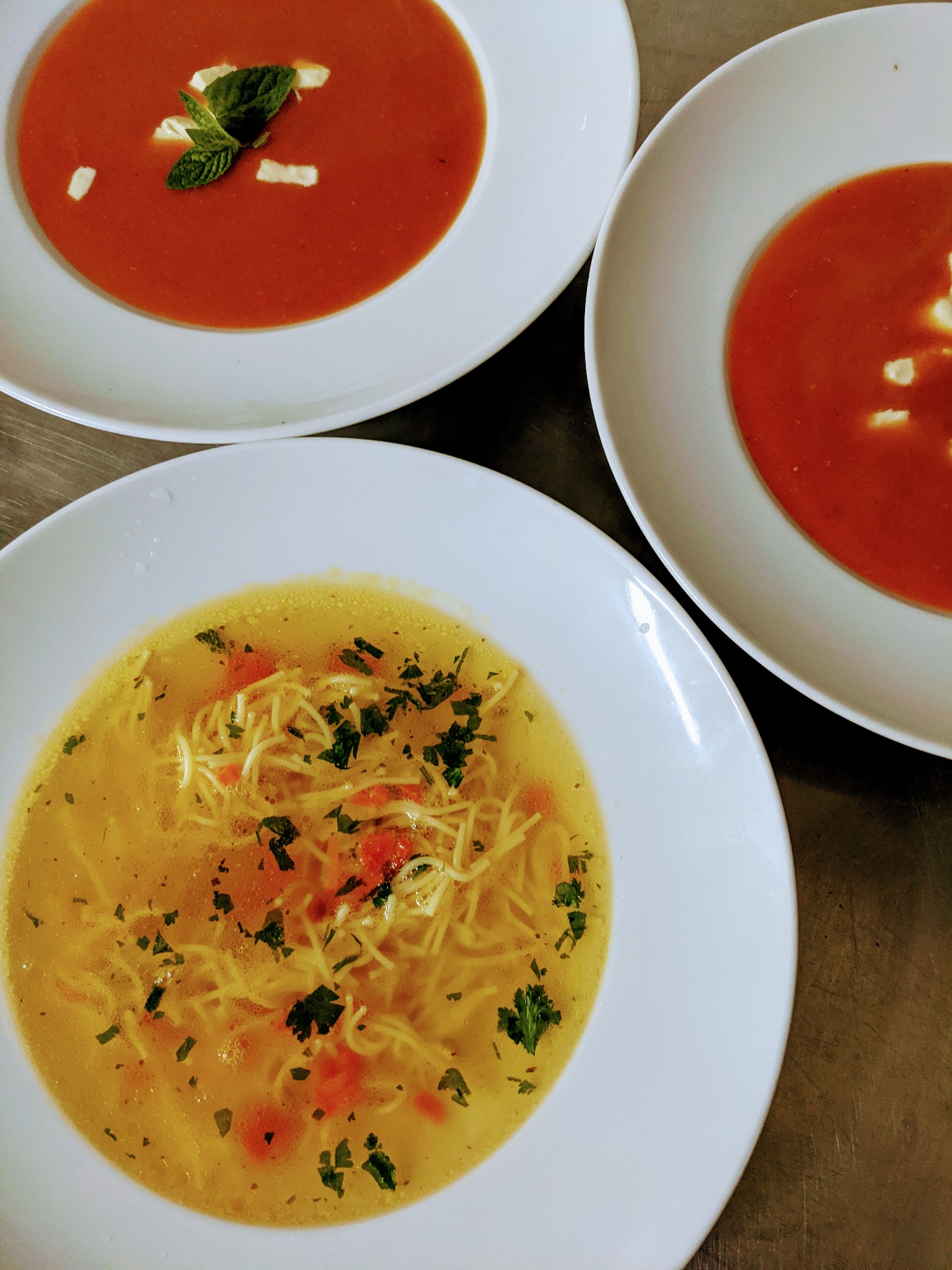 Trzy talerze z zupami: pomidorowa z listkami mięty i kostkami sera oraz rosół z makaronem i zieloną pietruszką, ujęcie z góry.