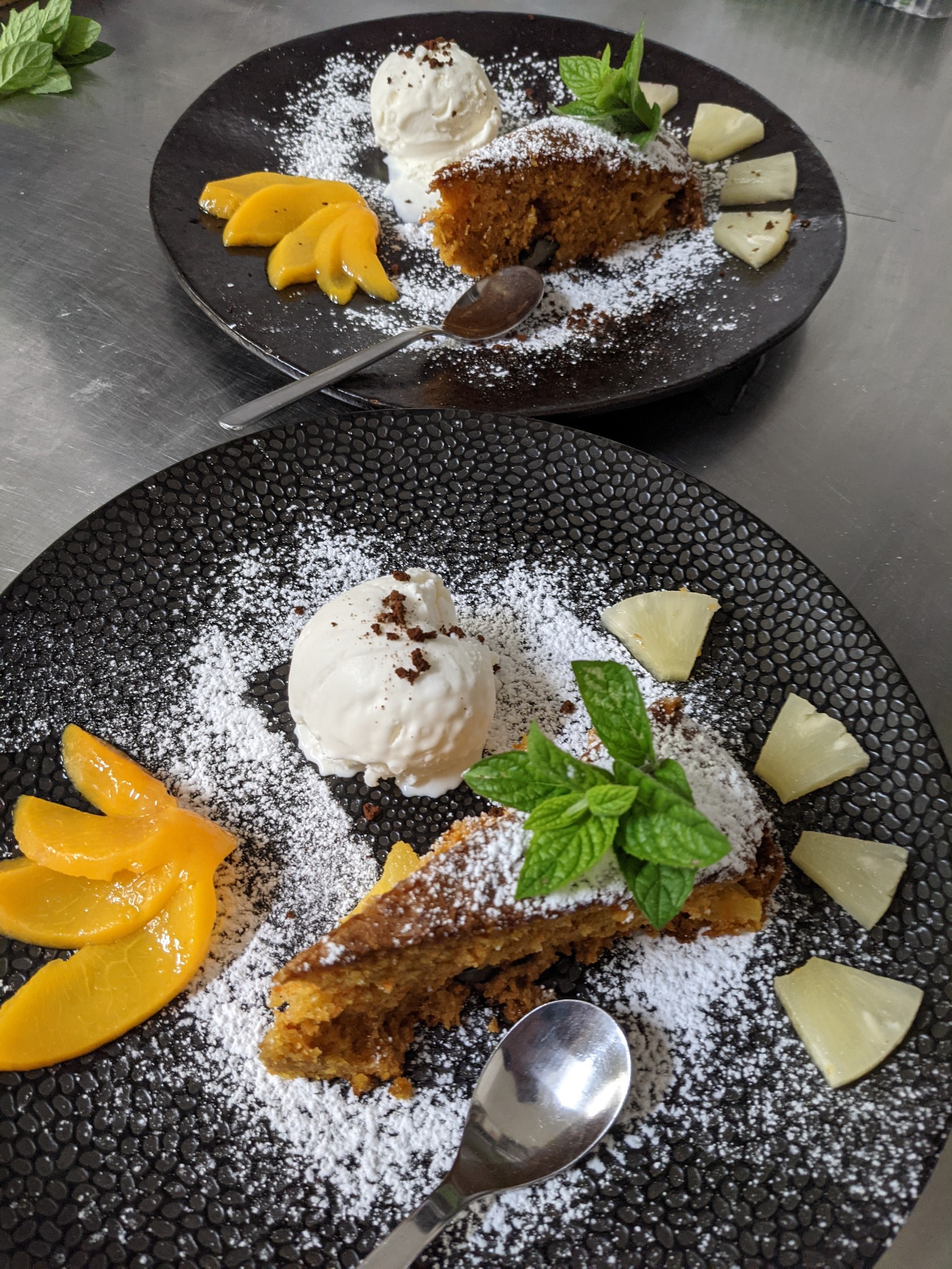 Dwa talerze z ciastem marchewkowym posypanym cukrem pudrem, gałką lodów, brzoskwiniami i ananasem, udekorowane miętą, na czarnych talerzach z teksturą.