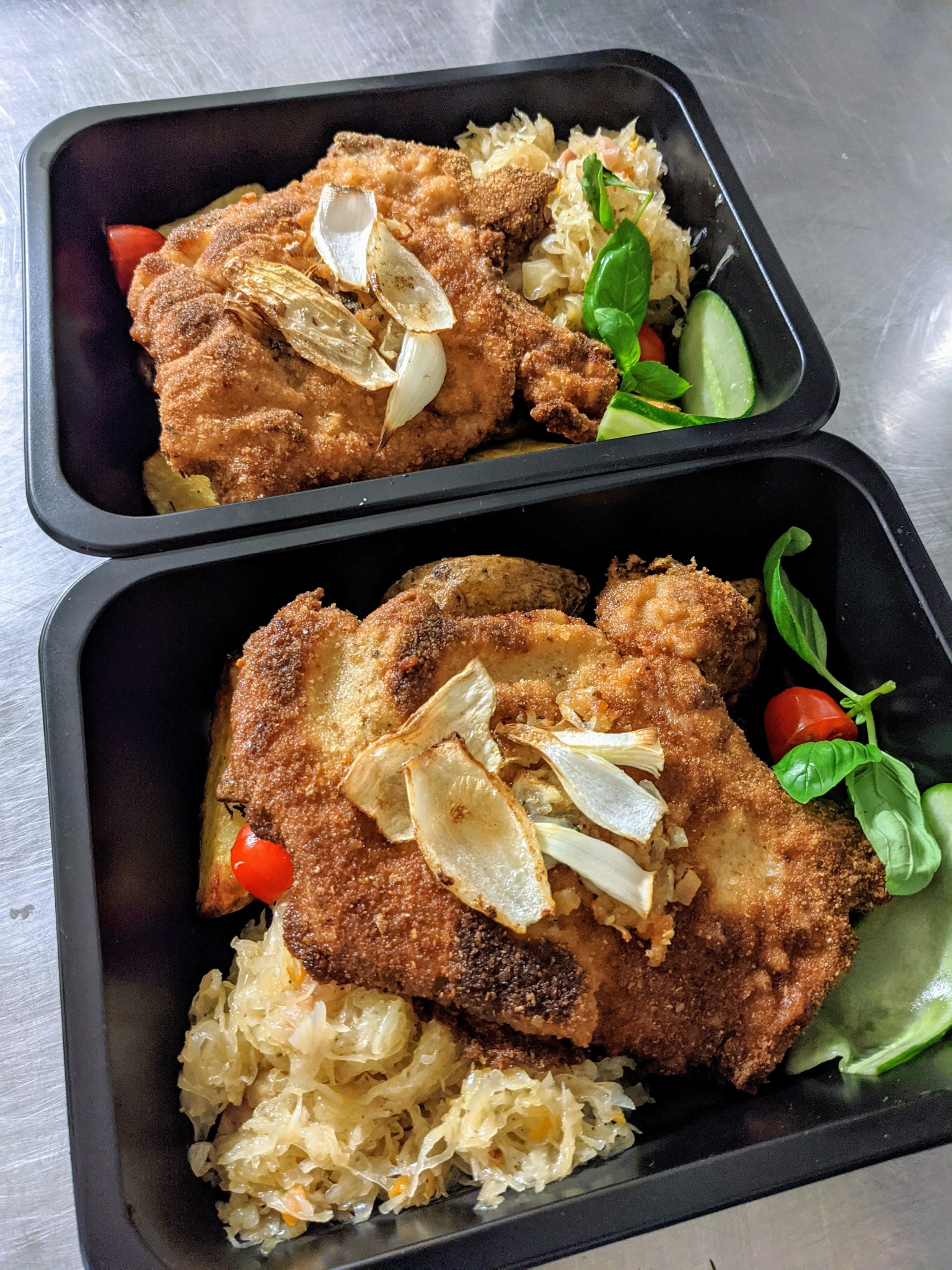 Dwa czarne pojemniki z gotowym daniem: panierowany kotlet schabowy z podsmażaną cebulą, kapustą kiszoną, pieczonymi ziemniakami, pomidorkami cherry, ogórkiem i bazylią, ułożone jeden na drugim.