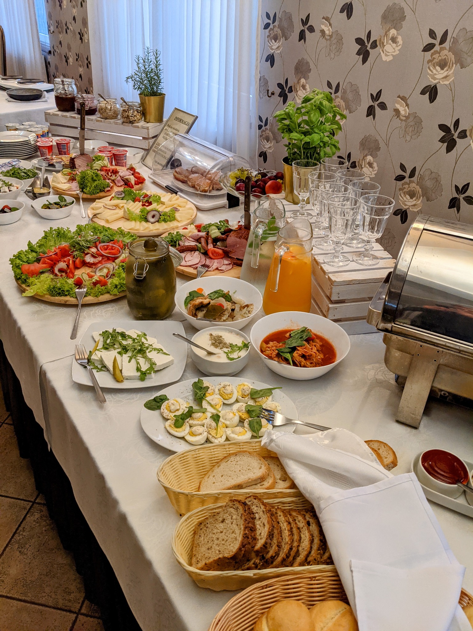 Bogato zastawiony stół szwedzki z różnorodnymi potrawami: sałatki, wędliny, sery, jajka, pieczywo, napoje i ciepłe dania, ustawione na białym obrusie w sali z dekoracyjną tapetą w kwiaty.