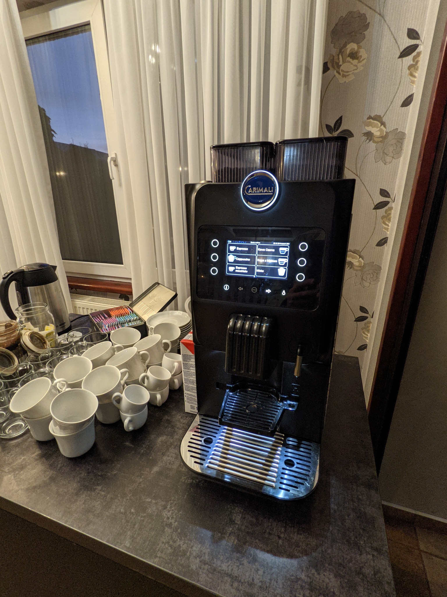 Profesjonalny, czarny ekspres do kawy Carimali z podświetlanym panelem dotykowym, otoczony stosem białych filiżanek i szklanek na ciemnym blacie, widoczny czajnik elektryczny i puszka z herbatami...