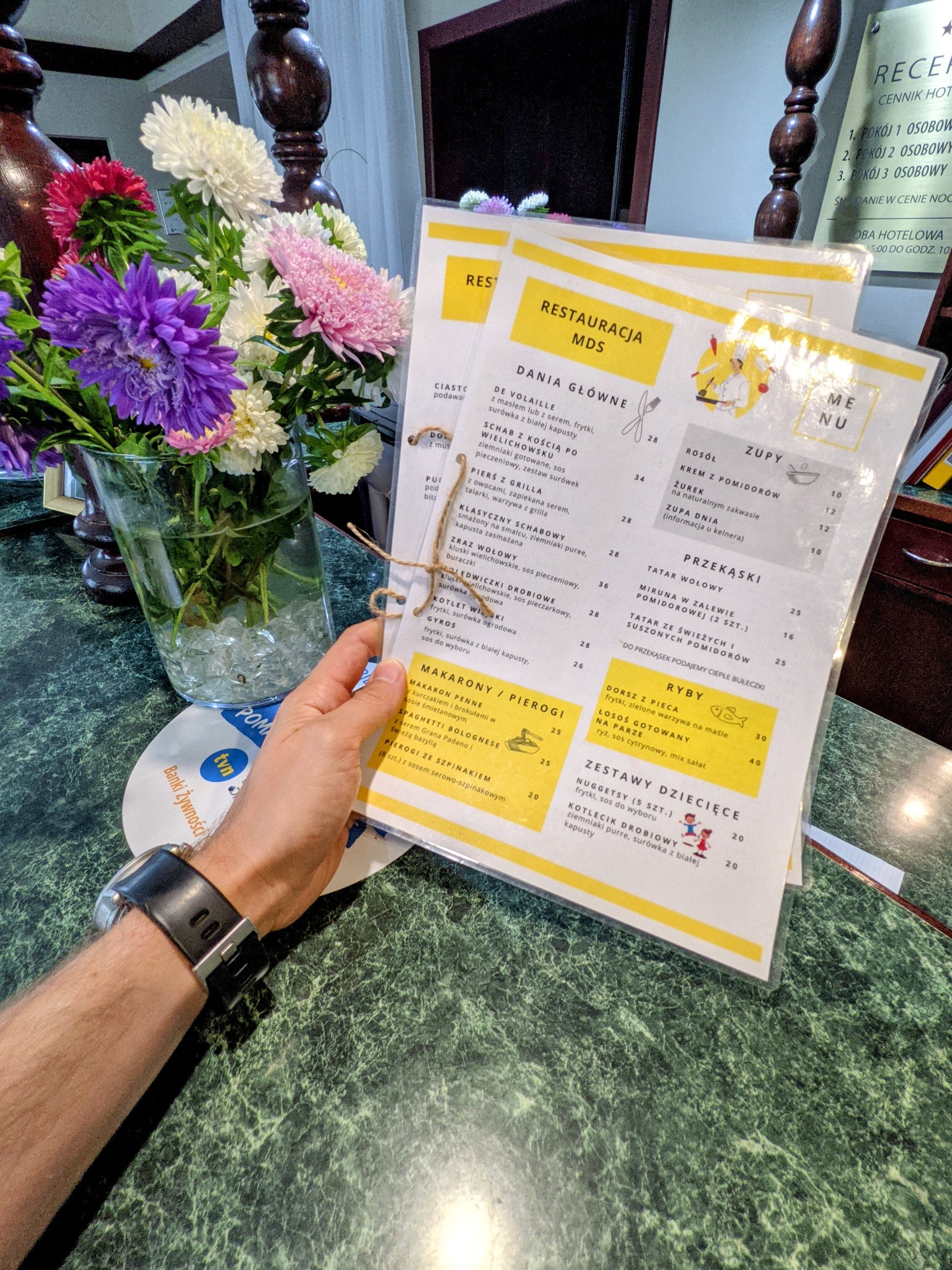 Menu restauracji 'Restauracja MDS' w przezroczystej okładce, trzymane w dłoni, z bukietem kolorowych astra w szklanym wazonie w tle, leżące na zielonym marmurowym blacie.