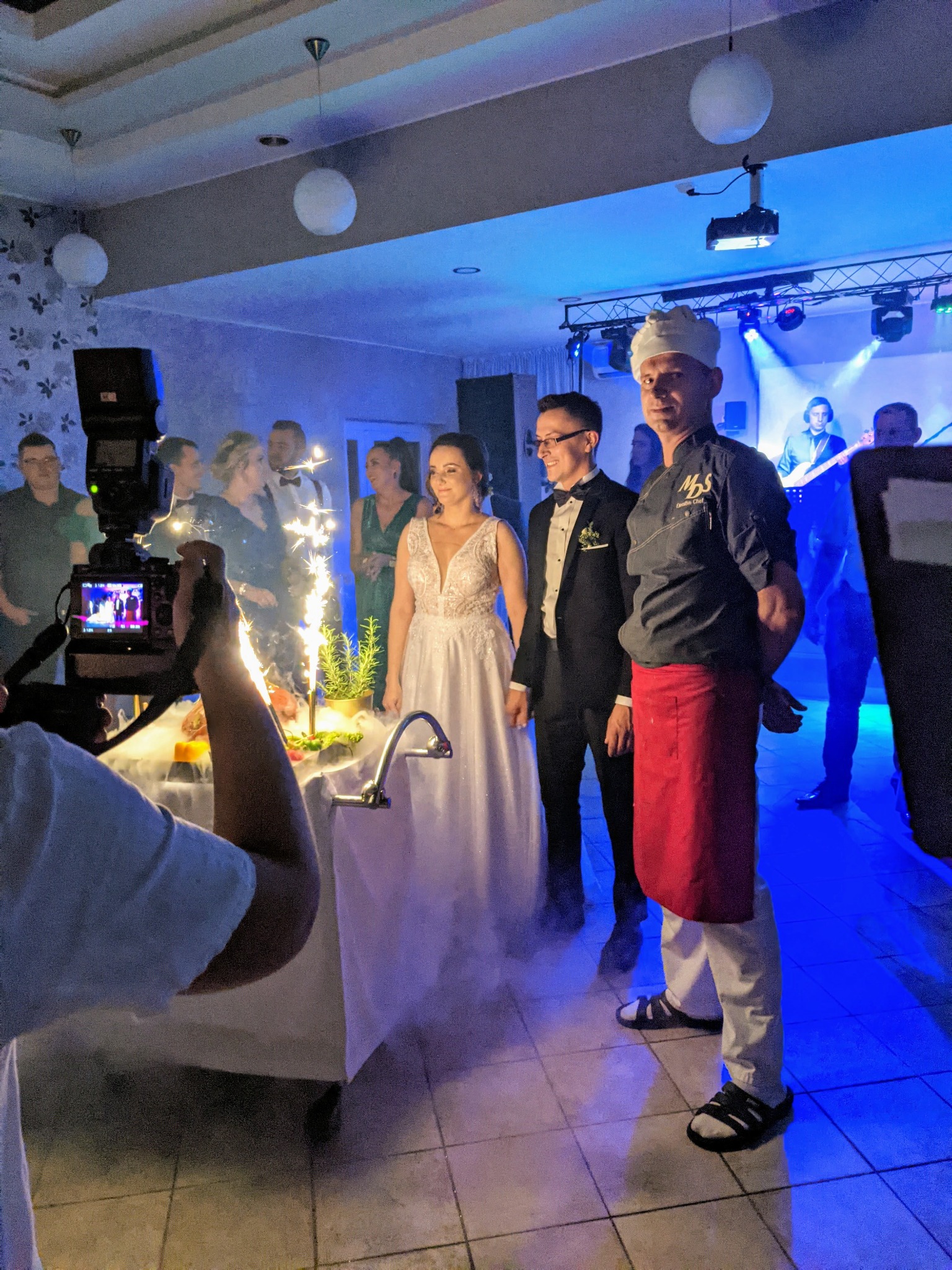 Uroczyste przyjęcie weselne: para młoda pozuje obok szefa kuchni w czapce i czerwonym fartuchu, w tle goście, zespół muzyczny na scenie i stół z dekoracjami i fontannami iskier, filmowane przez...