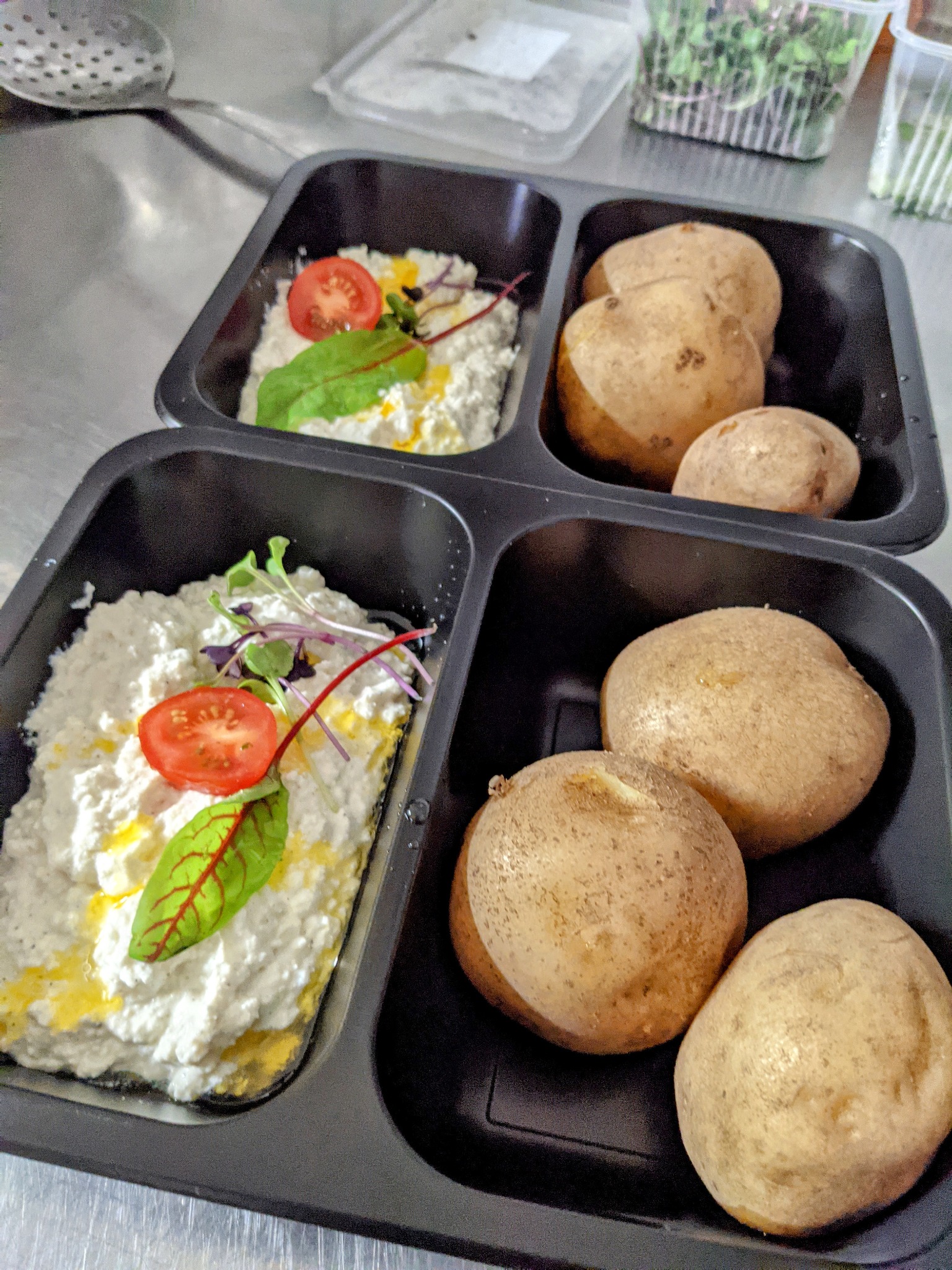 Gotowe danie dietetyczne w czarnym pudełku: twarożek z pomidorem i dekoracjami z zieleniny, obok ugotowane ziemniaki w łupinach.