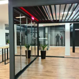 Nowoczesna, przeszklona konstrukcja z czarną ramą i lamelowym dachem z podświetleniem LED, widoczna na tle jasnego wnętrza z drewnianą podłogą i roślinami w donicach.