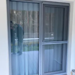 Przesuwne drzwi balkonowe z szarą ramą i moskitierą, widok z zewnątrz na taras z drewnianą podłogą, odbicie osoby w szybie.