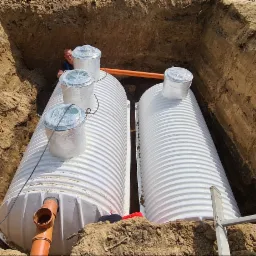 Instalacja dwóch białych, karbowanych zbiorników na ścieki w wykopie, widoczny pracownik wewnątrz wykopu, rury kanalizacyjne i drabina oparta o brzeg wykopu.