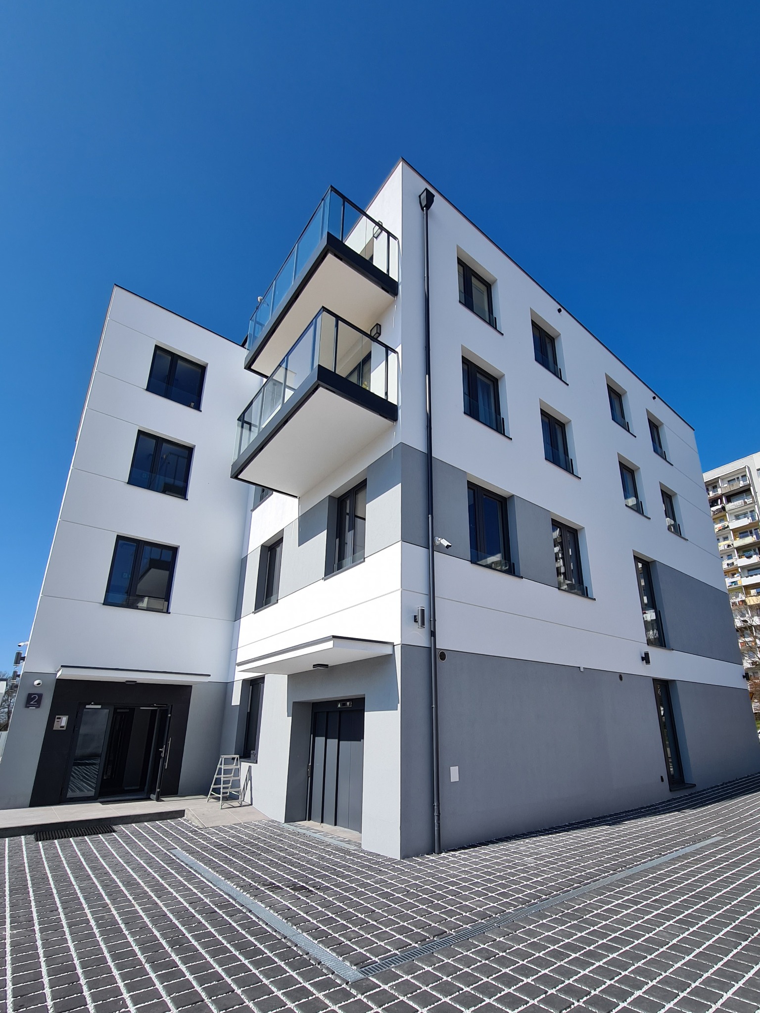 Nowoczesny budynek mieszkalny z balkonami o szklanych balustradach, czarnymi oknami PCV i elewacją w kolorze białym i szarym, widziany z perspektywy chodnika z kostki brukowej.