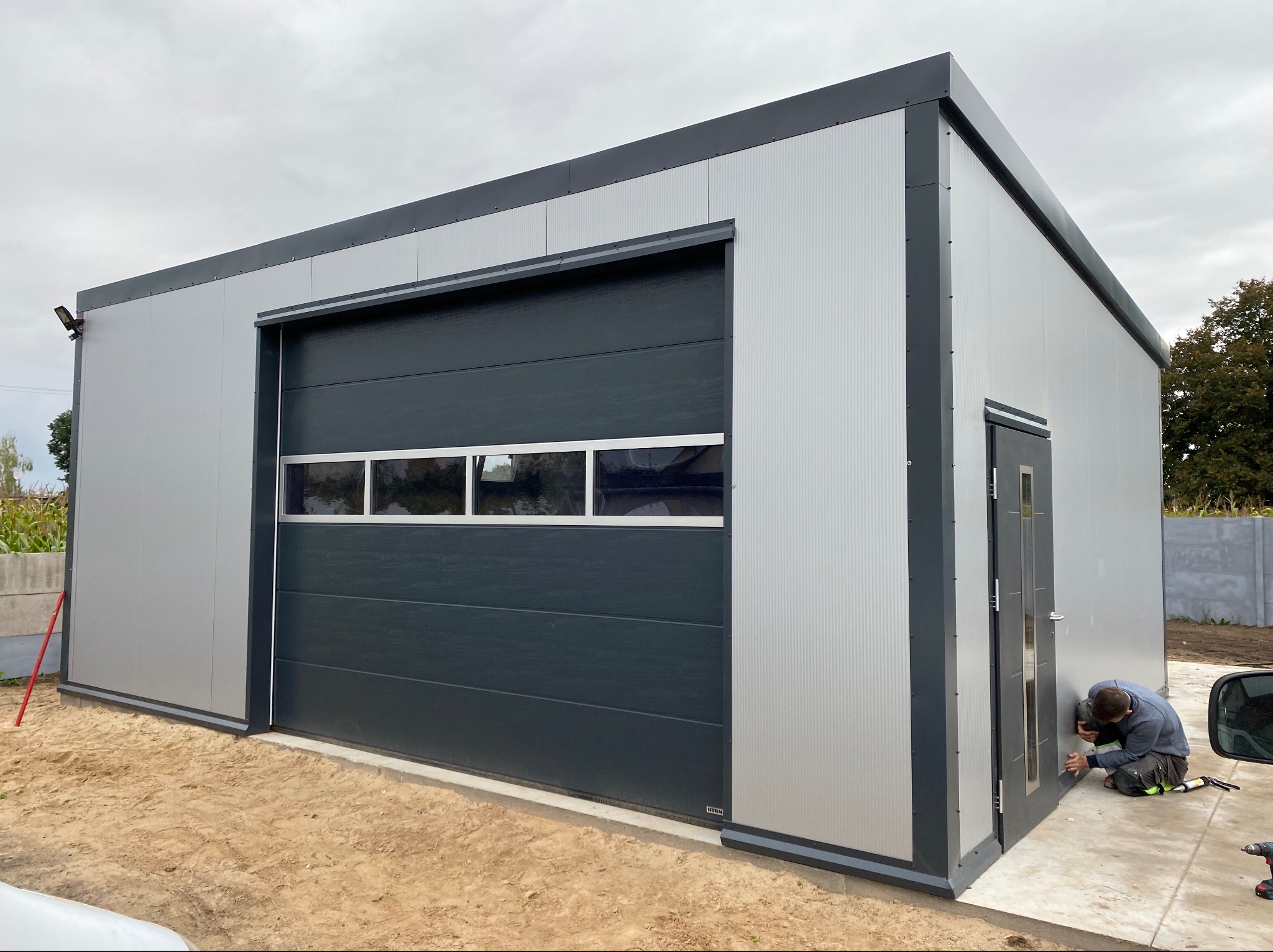 Nowoczesna hala z płyty warstwowej w odcieniach szarości, z segmentową bramą garażową z oknami, na etapie wykończenia, z pracownikiem montującym drzwi wejściowe.