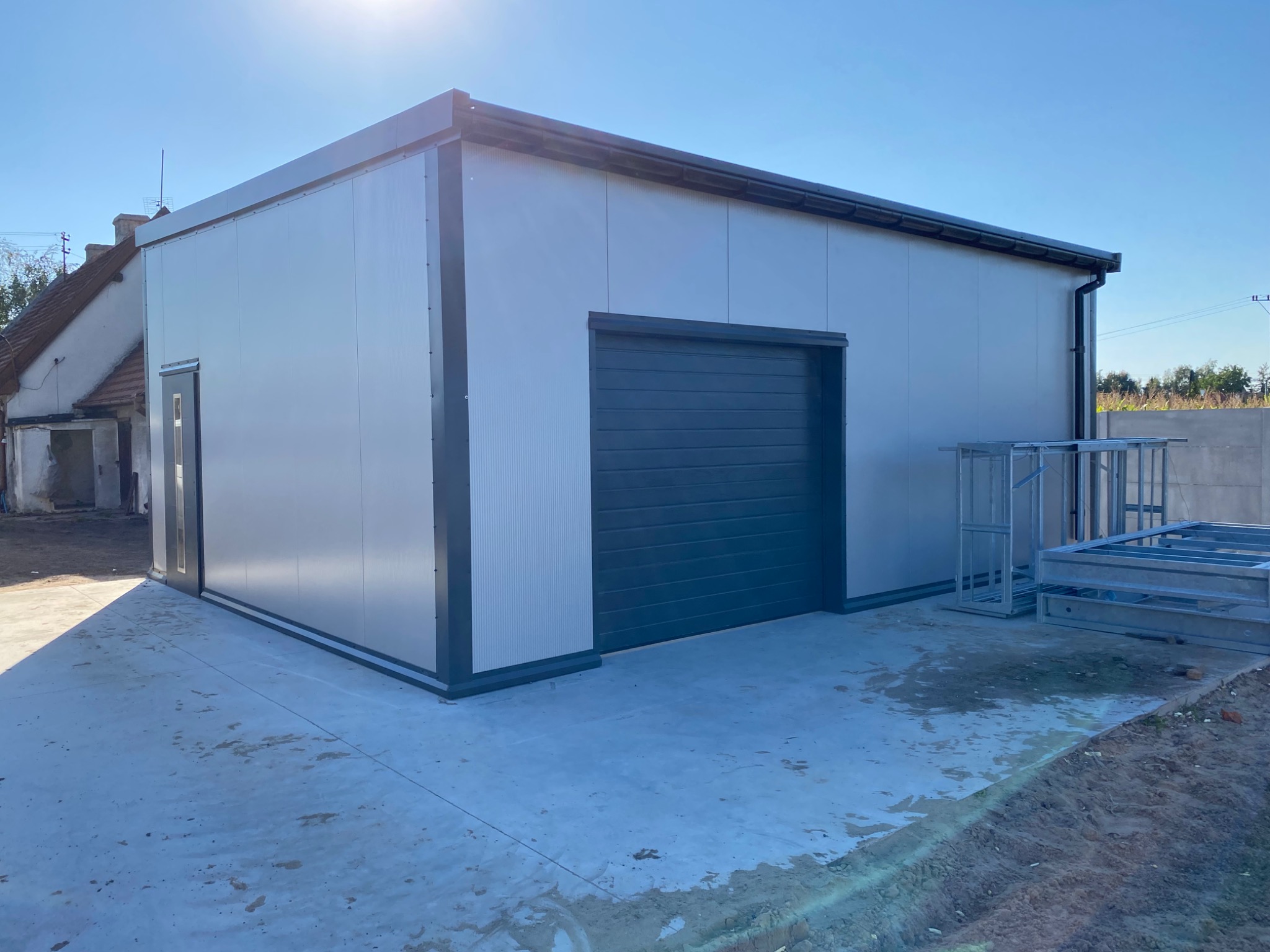 Nowoczesna hala z płyt warstwowych w jasnych kolorach, widoczna brama garażowa i drzwi wejściowe, na zewnątrz elementy konstrukcji stalowej.