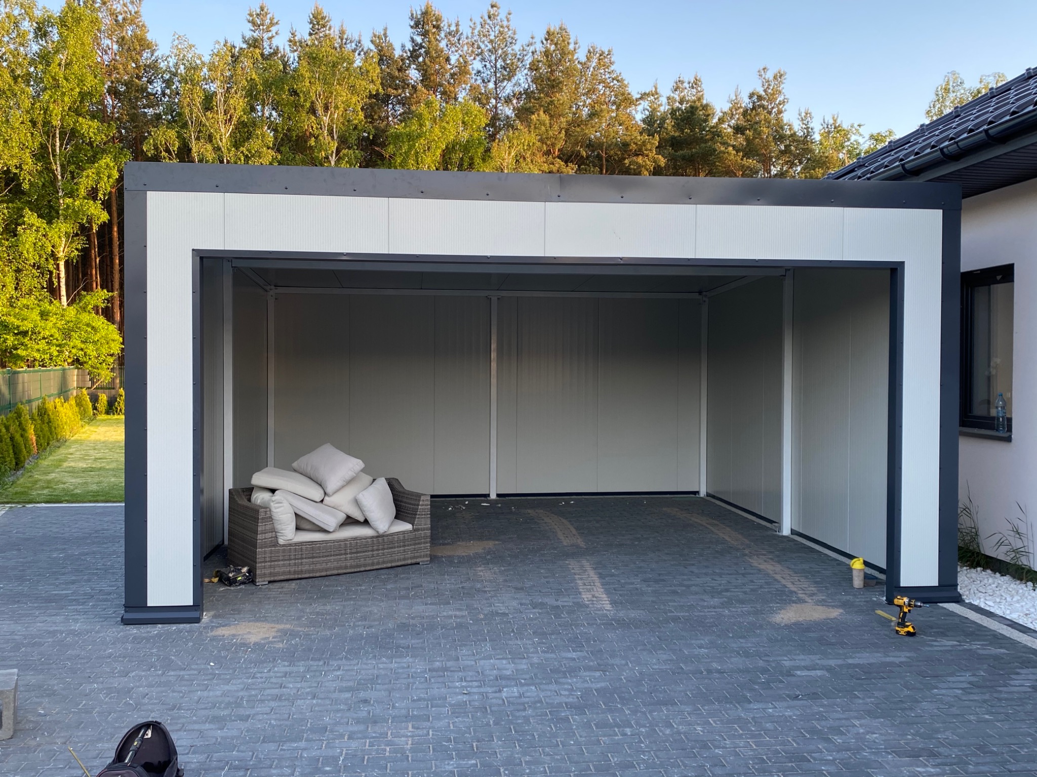 Nowoczesna, otwarta wiata garażowa z płyt warstwowych, wyposażona w meble ogrodowe i narzędzia, na tle domu jednorodzinnego i drzew.