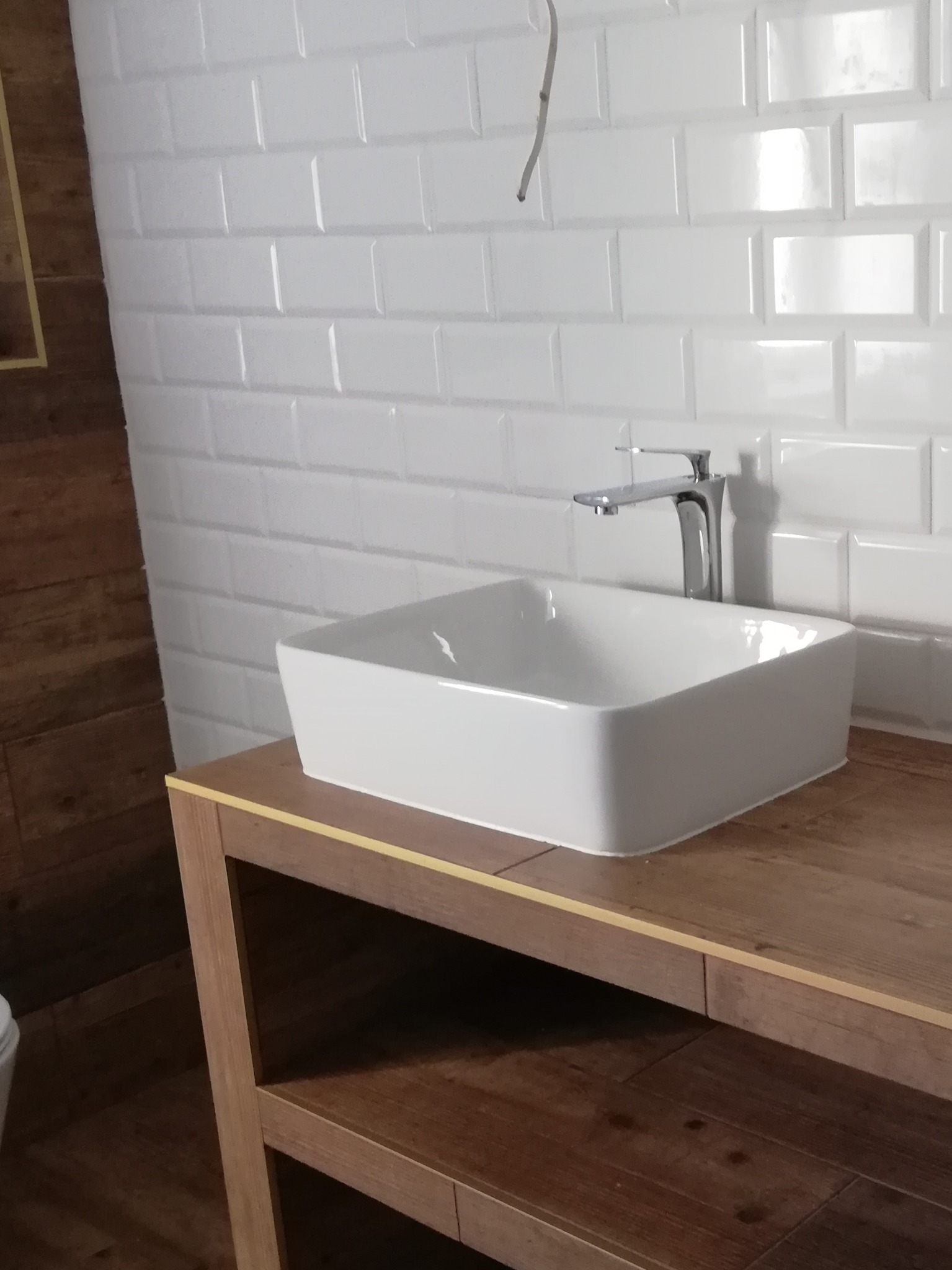 Biała, prostokątna umywalka nablatowa z chromowaną baterią, zamontowana na drewnianym, ażurowym blacie w łazience z białymi, błyszczącymi płytkami typu metro na ścianie.