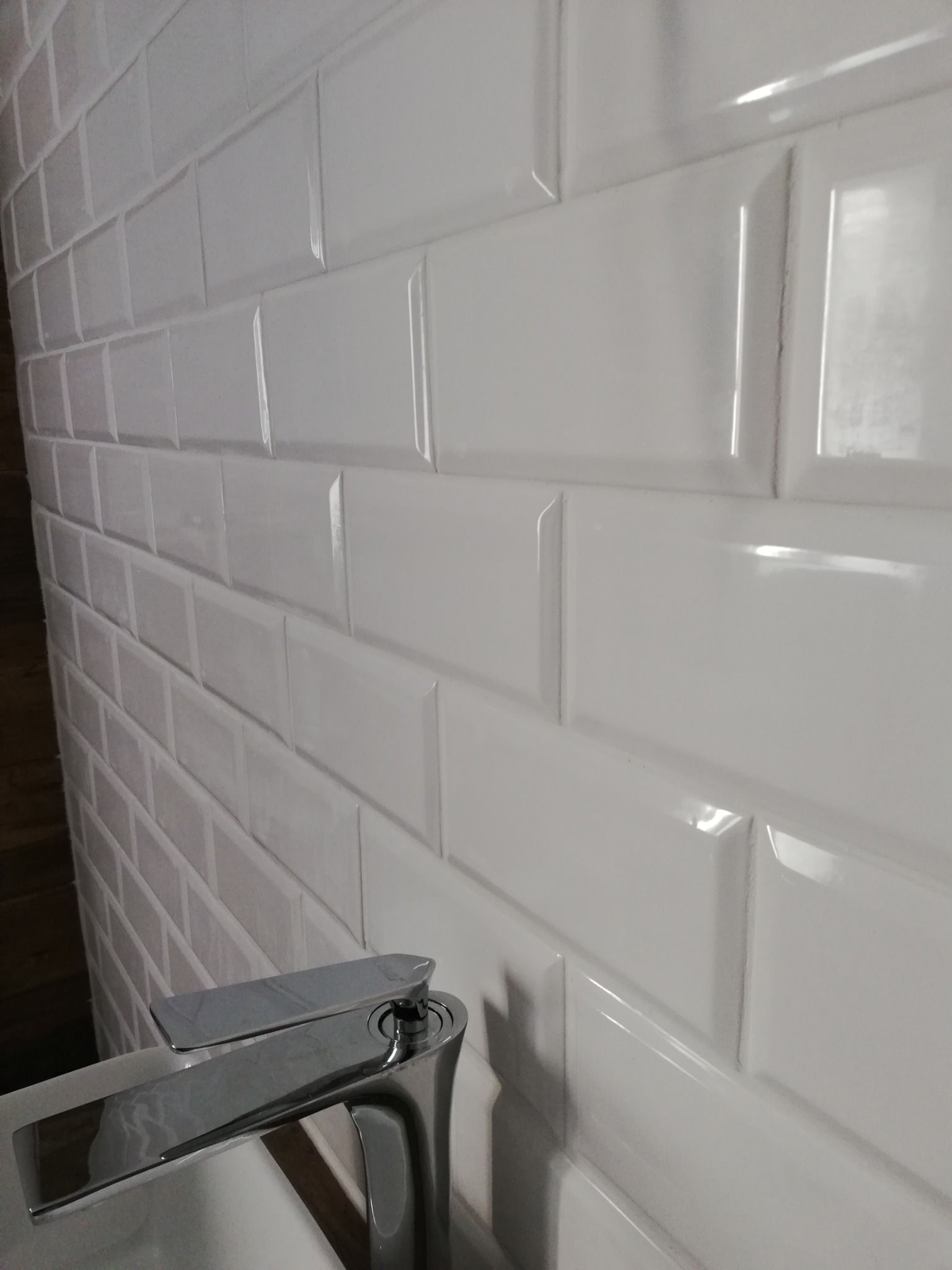 Białe, błyszczące płytki ścienne typu metro ułożone w łazience, widoczny fragment chromowanej baterii umywalkowej.