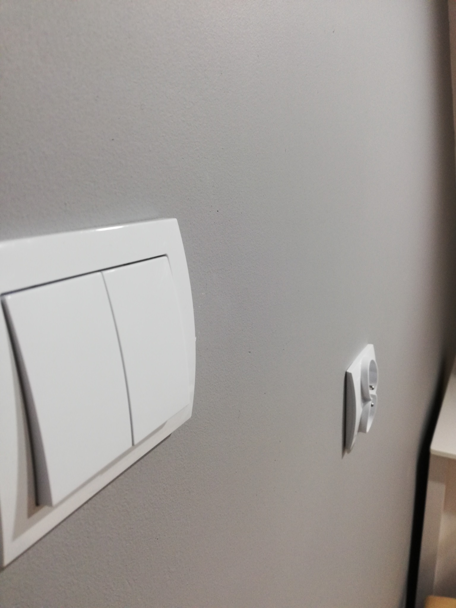 Szary pomalowany fragment ściany z zamontowanym białym gniazdkiem elektrycznym i białym podwójnym włącznikiem światła.