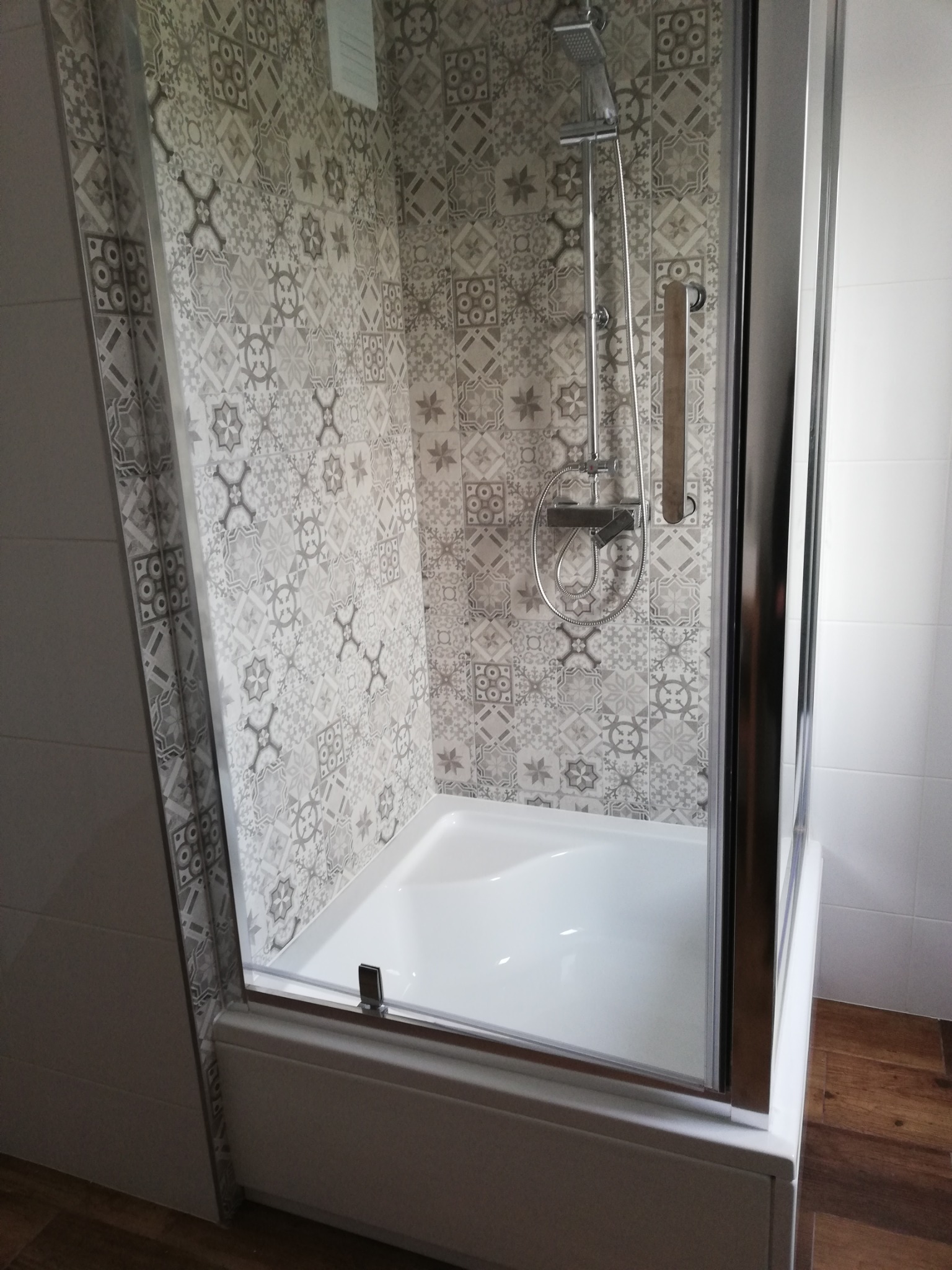 Szklana kabina prysznicowa z chromowanymi profilami, biały brodzik, ściany wyłożone płytkami w stylu patchwork w odcieniach szarości i bieli, widoczna bateria prysznicowa z deszczownicą.