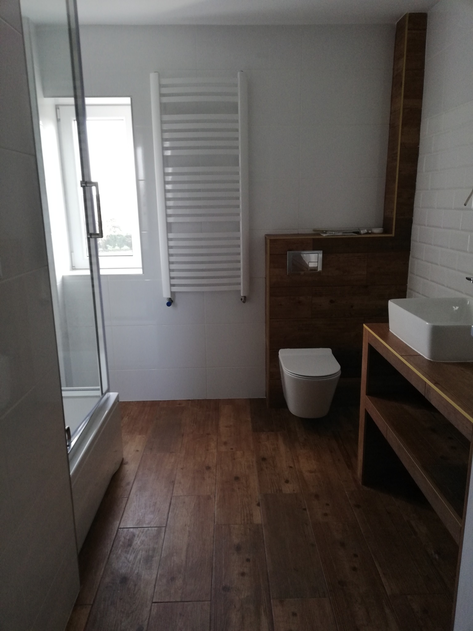 Nowoczesna łazienka z kabiną prysznicową, grzejnikiem drabinkowym, toaletą podwieszaną i umywalką nablatową na drewnianym blacie, wykończona płytkami imitującymi drewno na podłodze i częściowo...