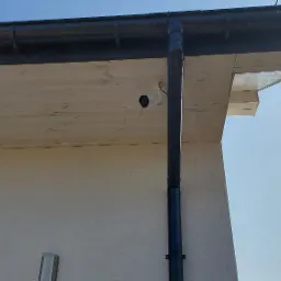 Biała kamera monitoringu Hikvision zamontowana pod drewnianym okapem, widoczna rynna spustowa, jasne tło ściany budynku i błękitne niebo.