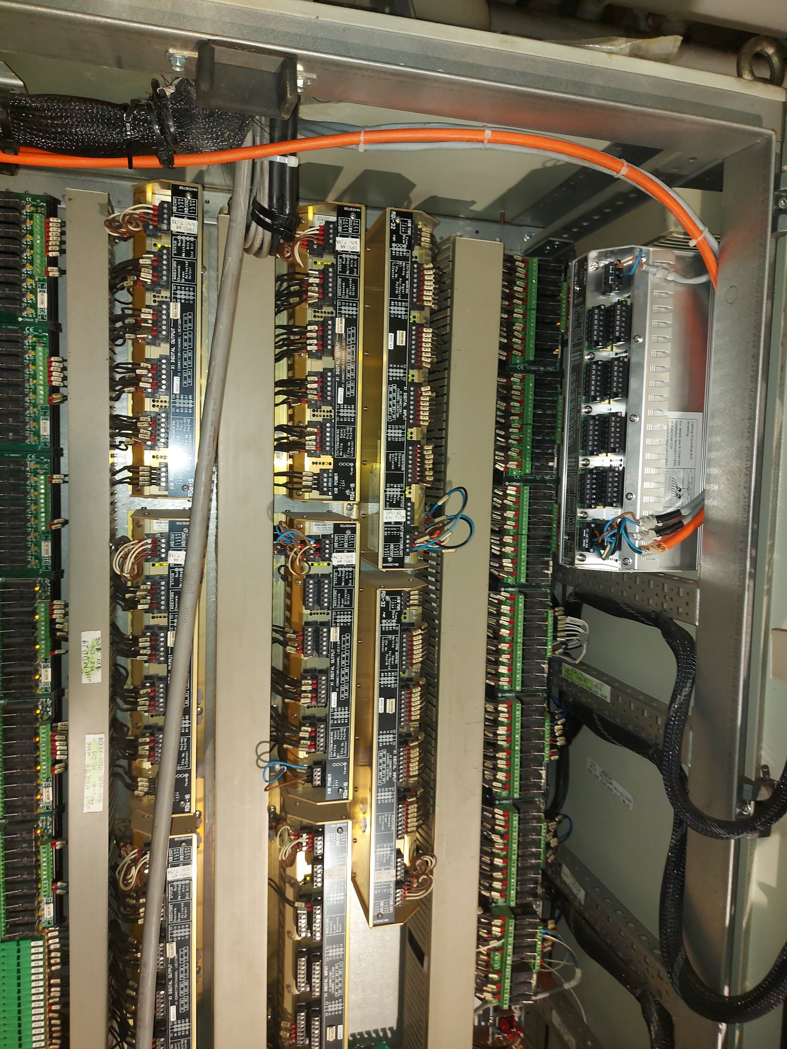 Wnętrze szafy sterowniczej z modułami wejść/wyjść, okablowaniem i elementami montażowymi, widoczne przewody w peszlach i oznaczenia na płytkach drukowanych.