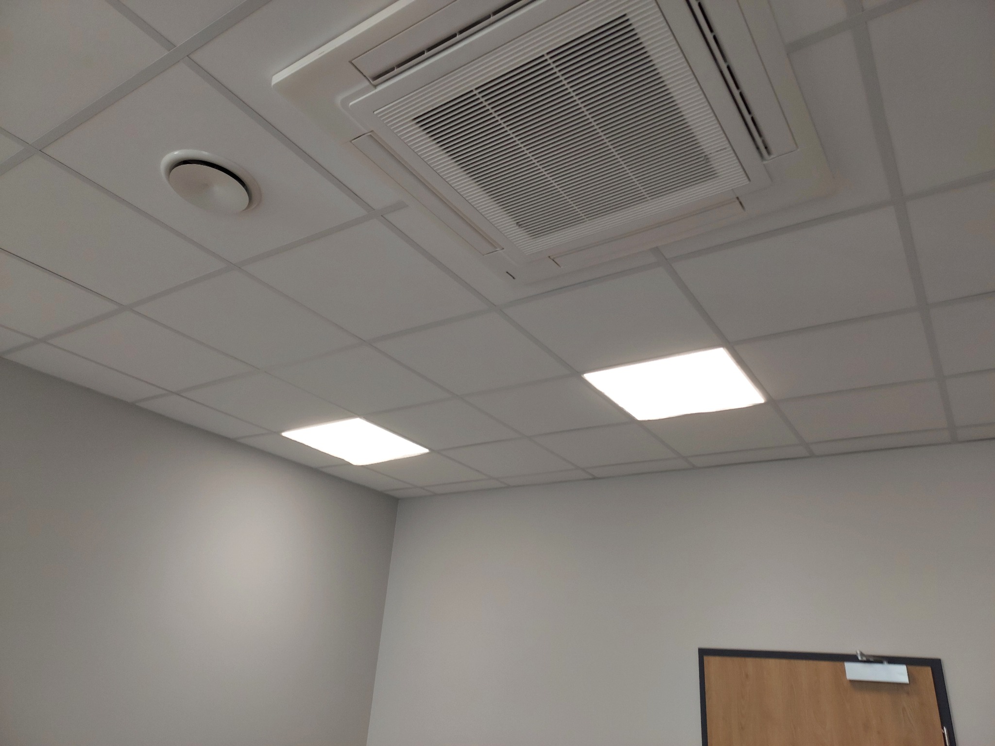 Sufit podwieszany z kratką wentylacyjną klimatyzacji kasetonowej, okrągły nawiewnik i kwadratowe panele oświetleniowe, w tle szara ściana i fragment drzwi.