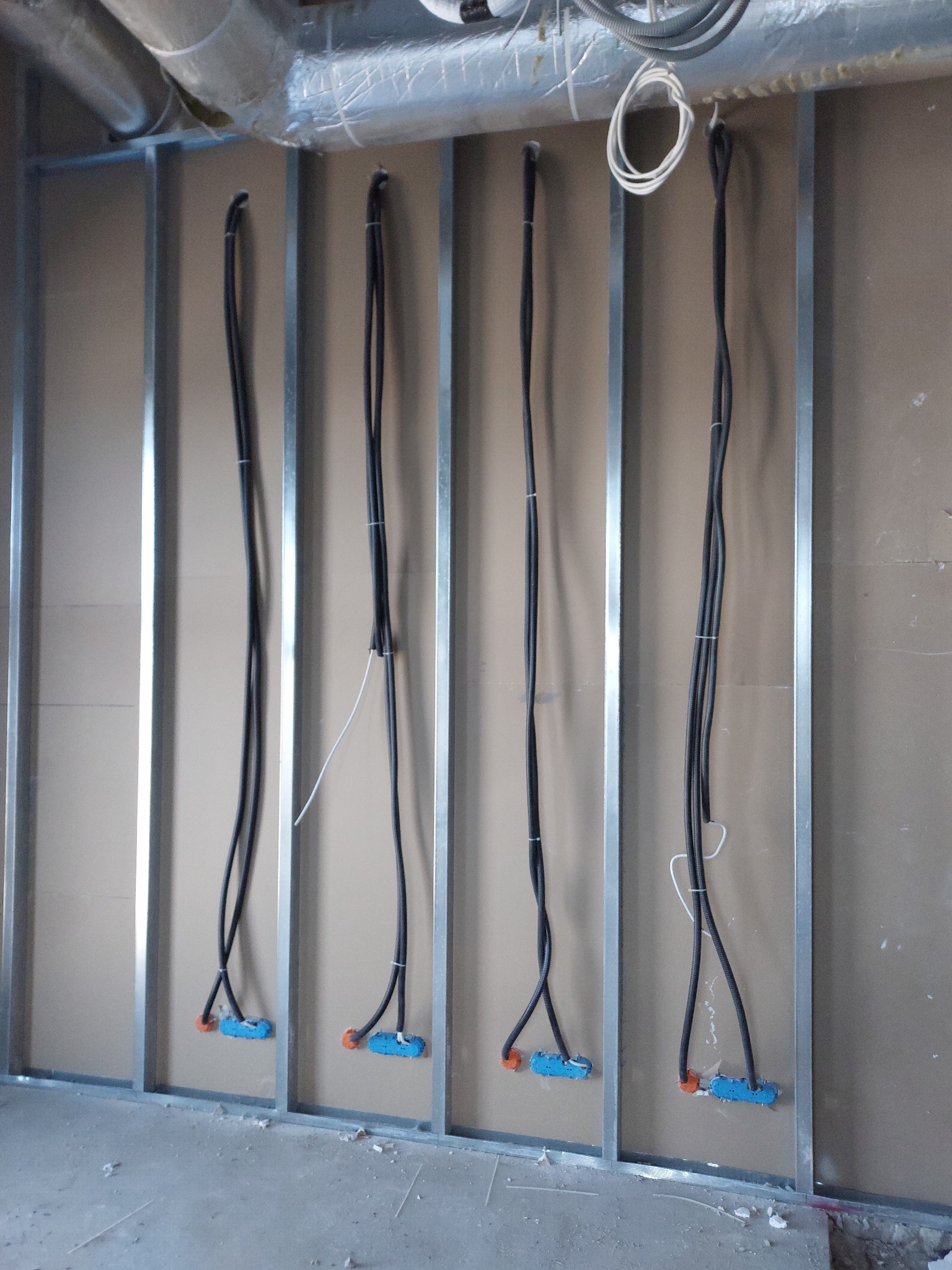 Cztery pionowe kable w peszlach, przymocowane do ściany z metalowych profili i płyt gipsowo-kartonowych, z niebieskimi puszkami instalacyjnymi u dołu.