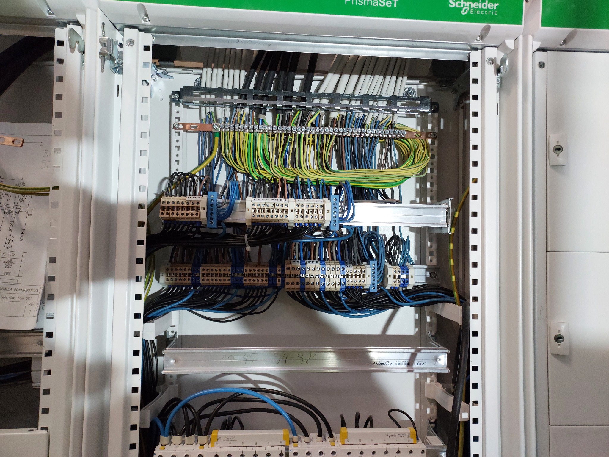 Rozdzielnica elektryczna z widocznymi przewodami w kolorach: czarnym, niebieskim, żółto-zielonym, podłączonymi do listew zaciskowych i zabezpieczeń, z etykietami producenta Schneider Electric.