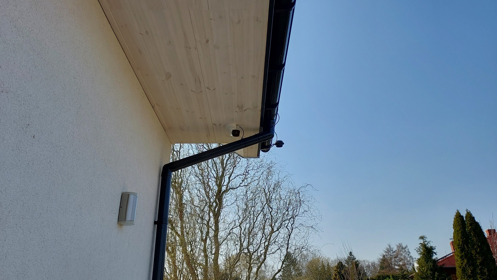 Dwie kamery monitoringu zamontowane pod okapem domu z drewnianą podbitką, widoczne orynnowanie i lampa zewnętrzna na elewacji.