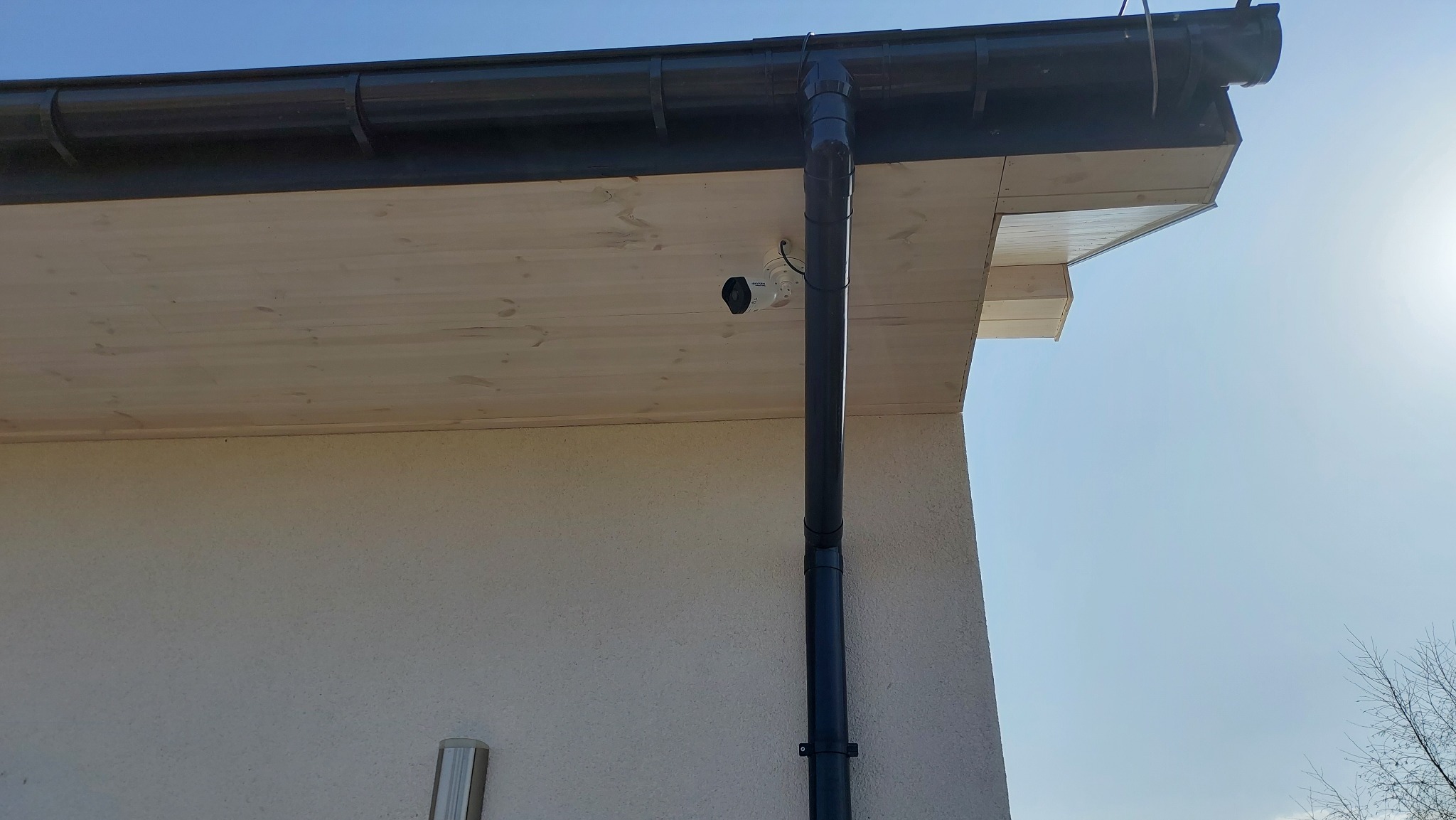 Biała kamera monitoringu Hikvision zamontowana pod drewnianym okapem, widoczna rynna spustowa, jasne tło ściany budynku i błękitne niebo.