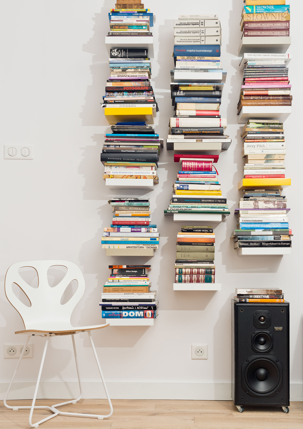 Białe, wiszące półki z książkami o architekturze i projektowaniu wnętrz na białej ścianie, minimalistyczne krzesło i czarny głośnik na drewnianej podłodze.