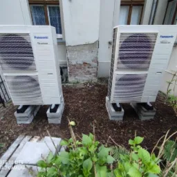 Dwa zewnętrzne moduły klimatyzacyjne Panasonic Inverter, ustawione na betonowych bloczkach przed budynkiem, z widoczną instalacją i roślinnością w tle.