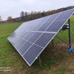 Panel słoneczny na metalowej konstrukcji, ustawiony na zielonym wzgórzu, widok na drzewa w tle, pochmurne niebo.