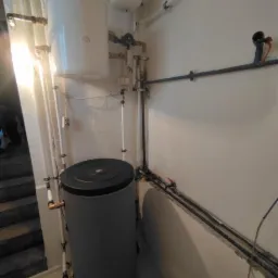 Szary zbiornik akumulacyjny ciepła z czarną pokrywą, biały zasobnik ciepłej wody i liczne rury instalacyjne na ścianie w pomieszczeniu technicznym.