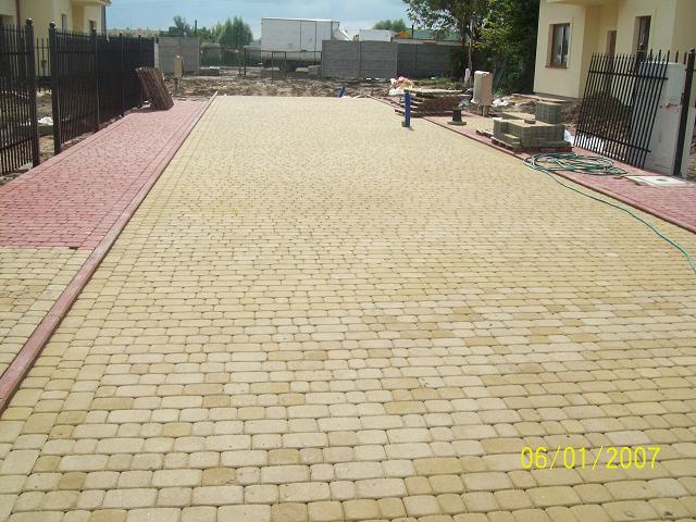 Ułożona nawierzchnia z kostki brukowej w odcieniach beżu i czerwieni, widok z góry, w tle budynki mieszkalne i ogrodzenie.