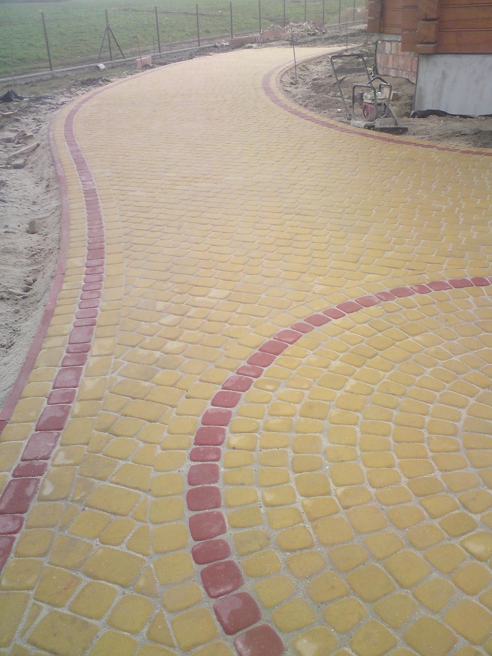 Ułożona ścieżka z kostki brukowej w odcieniach żółci i czerwieni, tworząca zakrzywiony wzór z okrągłym elementem na pierwszym planie, widoczna ubijarka w tle.