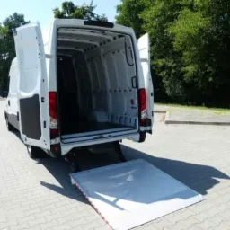 Biały samochód dostawczy z otwartymi tylnymi drzwiami i rozłożoną rampą załadunkową na brukowanym placu, w tle zielone drzewa.